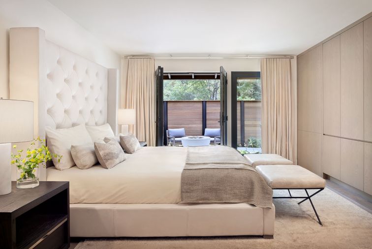 The master bedroom is a serene retreat overlooking a private patio, with a bed and nightstands from Restoration Hardware, Schumacher benches covered in Townsend Leather, and light-colored accessories from Room & Board, Village Interiors, Rubelli, C&C Milano, rug from Stacey Logan.