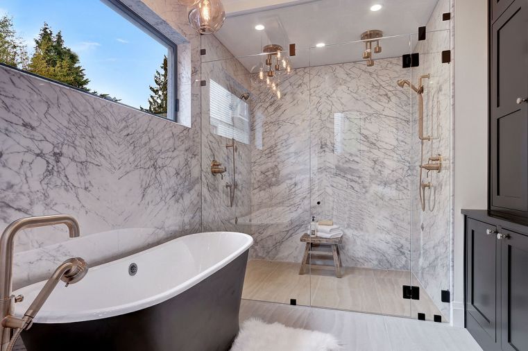 Master suite shower features dual Brizo luxe gold rain showerheads from the Litze collection. White Carrera honed marble walls extend throughout. Black matte exterior cast iron Kateryn Signature Hardware bathtub ties to shaker style cabinets at right.