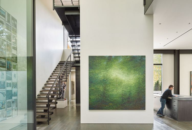 Lawrence Fodor (Friesen Gallery) oil on canvas entitled “Rain Forest” graces the entrance on a double-height foyer wall. The kitchen island is seen at right, while a sculpture features two businessmen fighting over money at left beneath the staircase rising to the second floor.