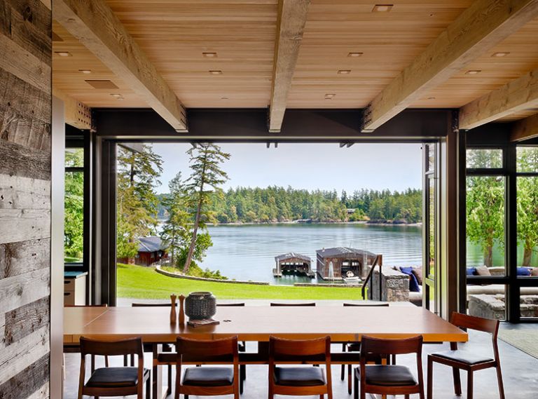 The new home, built to fit the vernacular of the historic compound, is situated on a knoll above great lawn looking toward Salish Sea through Quantum aluminum clad lift and slide doors. Custom dining table with stackable DWR profile chairs.