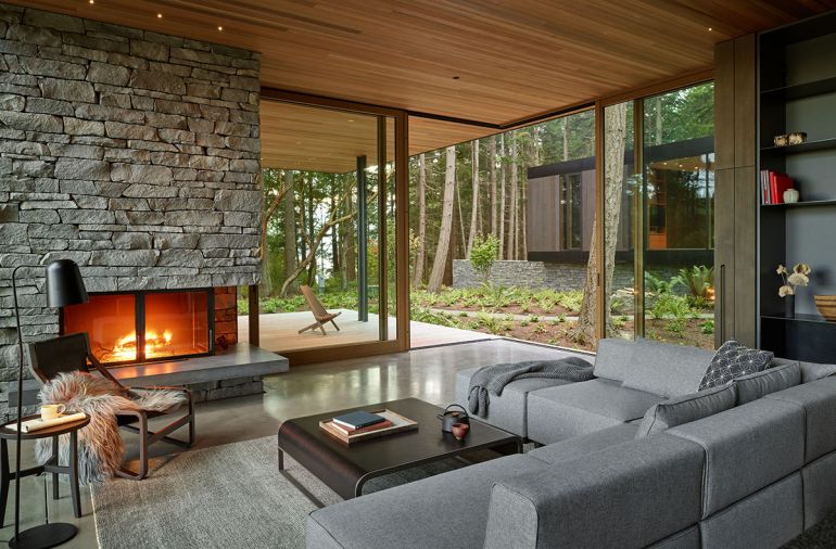 The corner of the living room opens to the Ipe deck and lush forest, and the Western Red Cedar ceilings extend outside. A sectional from Focus One Home and a Toro Lounge Chair by Blu-Dot are cozy spots by the Huckleberry basalt stone fireplace.