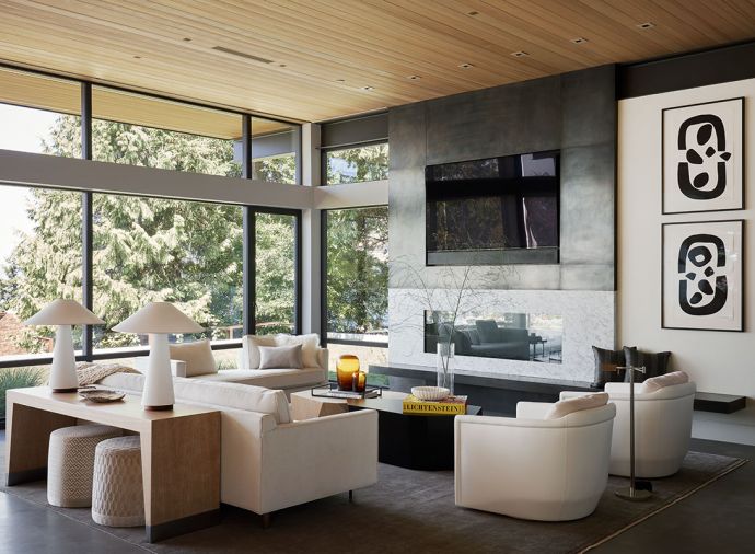 Fleetwood windows and doors were used throughout the home. The feature wall in the living room has custom blackened steel, marble, and a Da Vinci fireplace. A Mitchell Gold + Bob Williams sofa with Holly Hunt fabric sits across, with two Anees leather and mohair swivel chairs from Kelly Forslund at the Seattle Design Center. Poufs from Trammell-Gagne are tucked beneath the console, and the wall art is by Julie Speidel via Winston Wachter Gallery.