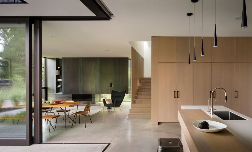 The kitchen is by Henrybuilt, utilizing plank-matched grey oak and Ravey Europly for the cabinetry. Ivory solid surface counters join a back-painted white glass backsplash. Micro Cone LED Pendant Light by Sonneman Lighting hangs above the island. The fireplace façade is formed from raw steel, and the stairs and screen are fashioned from grey oak. The floors are concrete.
