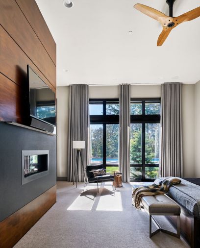 The relaxing master bedroom gets a simple set of window treatments; alder wood backed television with blued steel fireplace surround. Big Ass Haiku fan plays off Paul Schatz lamp, wooden stump side table, leather chair and bench.