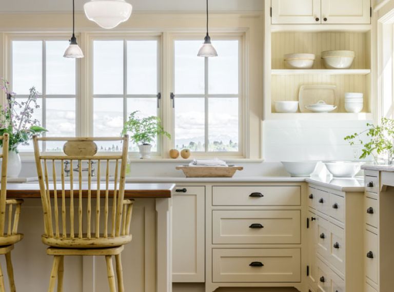 The traditional farmhouse kitchen is built around custom cabinetry from Warmington & North, a Seattle-based cabinetry specialist. Warm neutral tones on the walls, cabinets, area rug, and furniture enhance the glow that comes from wrap-around windows.