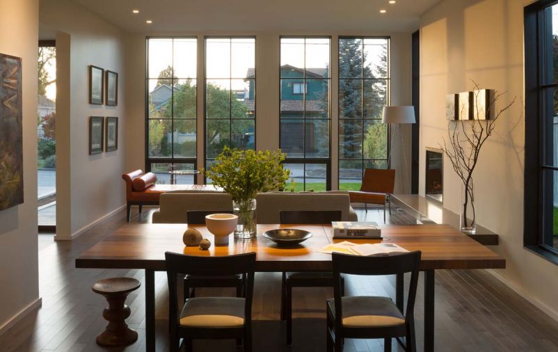 A crisp, clean-lined fireplace with a cantilevered honed quartz slab hearth anchors an open dining and living room layout. As the sun moves across the sky, shadows from the grid windows play across the fireplace wall.