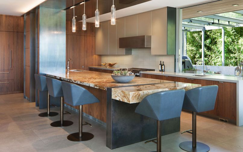 Handpicked polished granite countertop “Temptation” blends well with clear-stained black walnut cabinets. Blackened stainless steel left counters the back painted textured glass upper cabinet doors right. Kitchen back counter is Caesarstone – 3/4' quartz slab in Raw Concrete. Hexagon bar stools by Henge with Holly Hunt leather. Flooring is 2'x 4' grey Contrafalda porcelain tile. Handmade hanging pendant lights are part of the GCW lighting package. A pass-through window in the kitchen makes service to the BBQ area easy with a pocket window that slides behind the cabinetry.