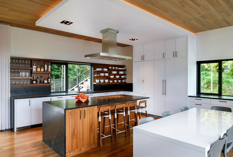 This kitchen was built for entertaining, with an open layout and plenty of space for cooking, hosting, and socializing. Dark soapstone counters contrast with white laminate cabinets and walnut accents, with oak flooring and a stained Western red cedar ceiling to match the exterior of the home. The oven, microwave, refrigerator, and dishwasher are from Miele, while the cooktop is from Monogram. A Blanco sink is finished with a KWC Luna faucet.