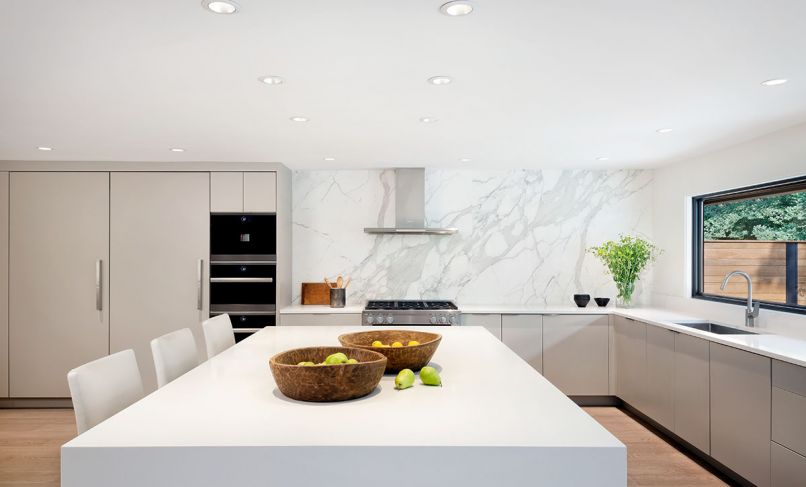 The pristine kitchen features vertical lines in the cabinetry, which is kept uncluttered thanks to tab pulls. Countertops from Pental Quartz are done in white, and a picture window above the sink overlooks the outdoor space.