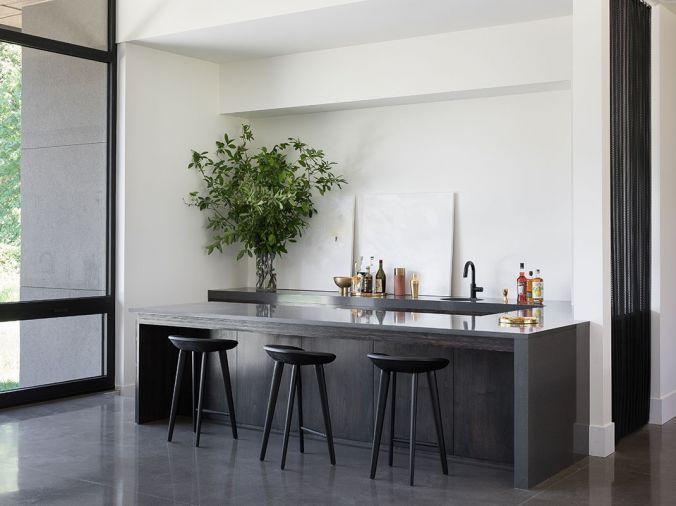 Radiant heat from ground source heat pumps that includes heat from the nearby pond keeps durable concrete floors running throughout the home cozy underfoot. The U-shaped bar features a stained, dark gray alder on the cabinet base with a dark gray MPP front and Pental quartz countertop. A curtain of raw steel coil by Cascade Home Décor separates the bar from kitchen.