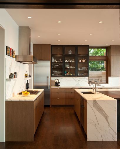 Upper black kitchen cabinet ties visually to the blackened steel entry piece and fireplace and recurs in the island’s footing. Waterfall marble island top countered by a Wolf Cooktop, Vent-a-Hood and repeating marble backsplash. Miele refrigerator is framed by kitchen cabinetry by Warner Cabinets. Photo © Rob Perry