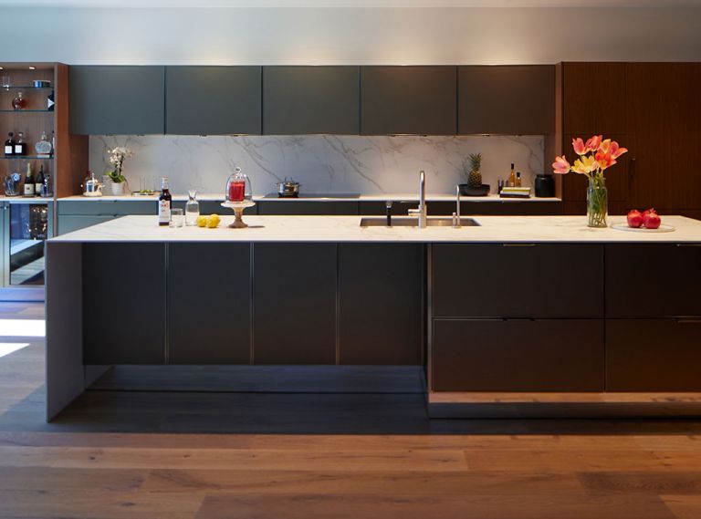 Grain matched walnut cabinetry is the main focus of the kitchen — used on the island and a bar area at one end of the kitchen and on the concealed refrigerator at the other, they are complemented by Gold Bronze upper cabinets, and Gold Bronze hardware and trim. All materials combine to complement each other and carry through the themes of minimalism and clean lines. Puck lights, mirrored toe kicks, and modern surfacing material used in the countertops and backsplash further emphasize the kitchen’s aesthetic while providing brightness and visual interest.