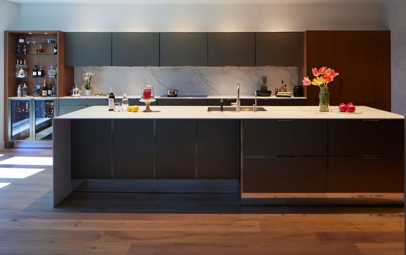 Grain matched walnut cabinetry is the main focus of the kitchen — used on the island and a bar area at one end of the kitchen and on the concealed refrigerator at the other, they are complemented by Gold Bronze upper cabinets, and Gold Bronze hardware and trim. All materials combine to complement each other and carry through the themes of minimalism and clean lines. Puck lights, mirrored toe kicks, and modern surfacing material used in the countertops and backsplash further emphasize the kitchen’s aesthetic while providing brightness and visual interest.