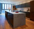 The lack of ornamentation in the kitchen allows the views of downtown Seattle to shine rather than compete with the space. In addition to matching the grain of the walnut cabinetry, the color seams running through the Dekton countertop and backsplash also align to provide a continuous visual flow. The homeowner intentionally chose a thin thickness of sintered stone to maintain the feeling of lightness. A cooktop from Gaggenau is barely visible and seems to float in the back counter. All other appliances in the kitchen are also from Gaggenau.