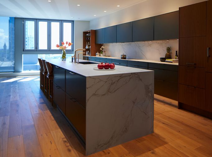 The lack of ornamentation in the kitchen allows the views of downtown Seattle to shine rather than compete with the space. In addition to matching the grain of the walnut cabinetry, the color seams running through the Dekton countertop and backsplash also align to provide a continuous visual flow. The homeowner intentionally chose a thin thickness of sintered stone to maintain the feeling of lightness. A cooktop from Gaggenau is barely visible and seems to float in the back counter. All other appliances in the kitchen are also from Gaggenau.