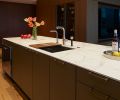 In the foreground, a sink from the Julien Smart Sink collection and a faucet from Dornbracht are set into the kitchen island. Due to the openness of the kitchen and living space, the homeowner asked Cathleen Summers to design a cabinetry transition between the two areas. In response, she created a multi-use wall of walnut that houses a pantry and other kitchen storage behind panels and doors while the side fronting the living space holds the television and hides media equipment, home office supplies and other home trappings from view.