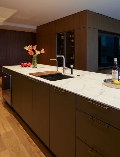 In the foreground, a sink from the Julien Smart Sink collection and a faucet from Dornbracht are set into the kitchen island. Due to the openness of the kitchen and living space, the homeowner asked Cathleen Summers to design a cabinetry transition between the two areas. In response, she created a multi-use wall of walnut that houses a pantry and other kitchen storage behind panels and doors while the side fronting the living space holds the television and hides media equipment, home office supplies and other home trappings from view.