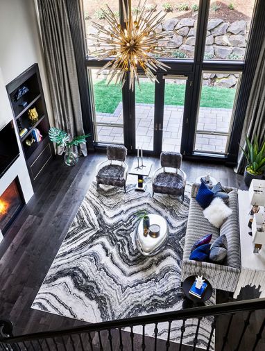 The view from the central staircase highlights the updated fireplace with floor-to-ceiling chimney and dark wood built-ins. Sherwin Williams Caviar painted window trim and molding adds drama. Hudson Valley chandelier provides glam. Feizy rug underscores modern design. Bernhardt sofa with Art Deco velvet upholstery and furry chairs give comfort.