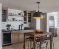 To create continuity, the main floor features a sandy-colored, weathered-wood-like flooring that extends throughout the space, including into a breakfast bar and beverage area adjacent to the kitchen. Here, dark gray granite countertops are flecked with shimmer, and the shape and finish of the light fixture relates to the gold trim on the bar island. Cabinetry is maple with a pumice finish, which nods the stone tile material that clads the home’s fireplace.