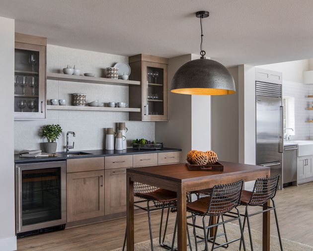 To create continuity, the main floor features a sandy-colored, weathered-wood-like flooring that extends throughout the space, including into a breakfast bar and beverage area adjacent to the kitchen. Here, dark gray granite countertops are flecked with shimmer, and the shape and finish of the light fixture relates to the gold trim on the bar island. Cabinetry is maple with a pumice finish, which nods the stone tile material that clads the home’s fireplace.