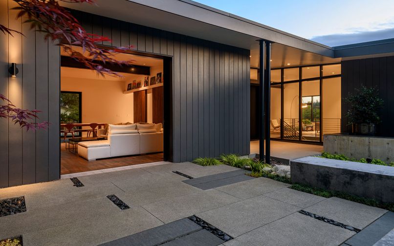 An Acer Emperor maple graces the courtyard looking through the Weiland sliding door into family room. Patio landscape design with poured-in-place 2x4 concrete pavers interrupted by thermalled basalt pavers countered with polished black pebbles. L-shaped cement bench backed by crème de mint Pittosporum. Living room seen through Quantum aluminum clad windows beyond.