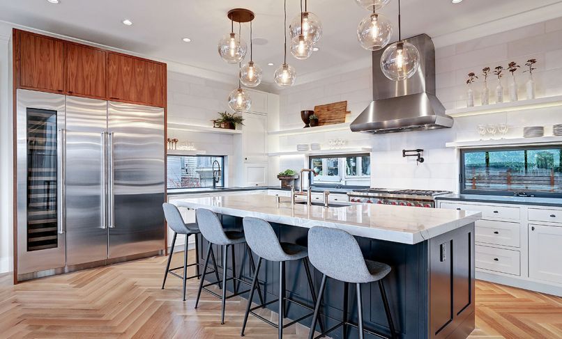 Sub-Zero trio of beverage cooler, freezer and refrigerator is crowned by warm American walnut cabinetry that adds visual contrast to white oak herringbone flooring. Three West Elm Sculptural Glass pendants in Champagne provide task lighting over island with Mont Blanc quartzite countertop. Free-hanging shelves painted to match shaker cabinets below add dimension to the Fleetwood awning windows that flank the Wolf range and the 8' x 8' Empyrean Ice wall tile. Convenient pot filler on range backsplash.