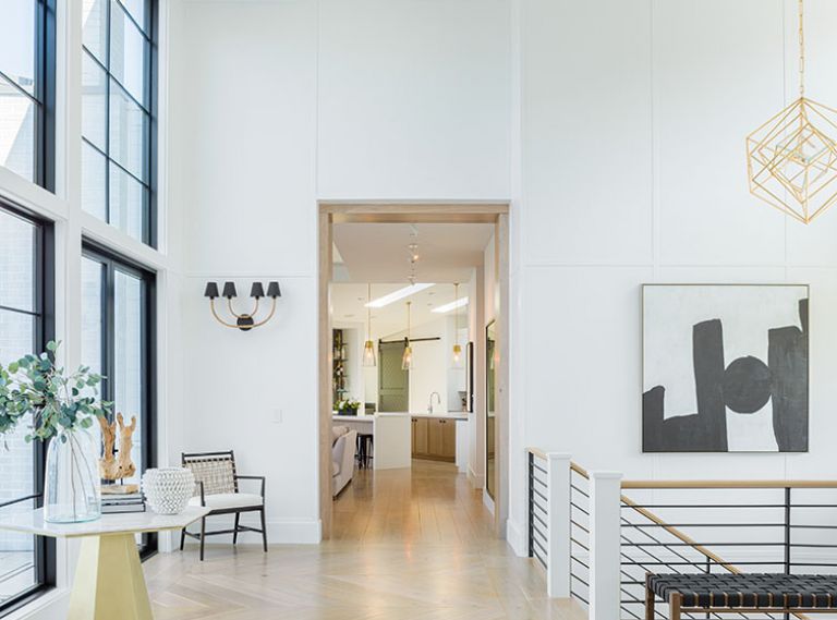 Dumont reiterates black, white, and gold palette throughout entry: painted wood paneling, steel staircase, Visual Comfort Cubist pendant, Arteriors sconces, hex-shaped table, and artwork. French doors flank Marvin aluminum clad windows.