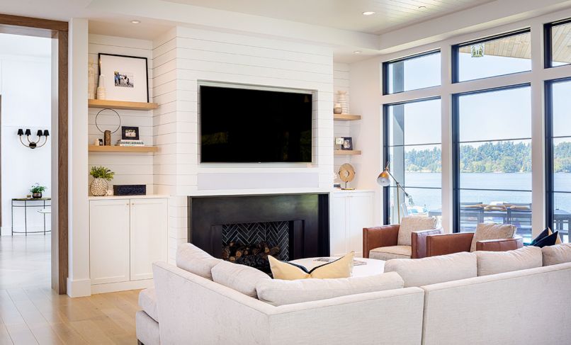 Dumont laid out the custom stained tongue and groove chevron wood flooring in entry by Salisbury Woodwork of Bainbridge Island. Shiplap wall and TV surround contrasts with waxed blackened steel fireplace and black indigo herringbone stone insert. J Redmond sectional with stone fabric faces off with pair of cognac Dorado leather-trimmed chairs from J Garner Home. White oak stained shelving echoes kitchen cabinetry. Marvin windows open to water view. Sturman’s custom walnut trimmed archways are an eye-catching detail.