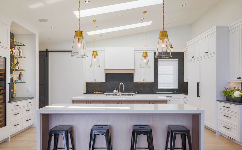 Dual islands feature waterfall Pental quartz countertops. Woodway Woodwork cabinetry with Ashley Norton hardware. Dumont and homeowner chose unique black porcelain slab from Pental as backsplash and perimeter countertops. Gold bracketed French bistro glass shelving adds contrast. Visual Comfort Robinson gold pendants.