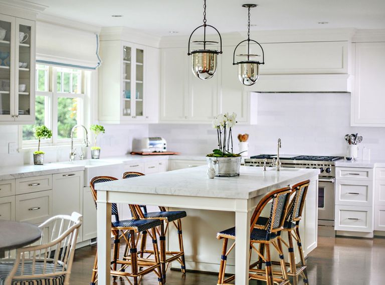 The renewed kitchen was taken from Tuscany to Nantucket with the use of white cabinets, counters and backsplash. The change answered the homeowners’ desires for a light-filled space that honored the home’s exterior architecture. To marry old and new and here and there, the designers used statuary marble on counters, Parisian stools are placed around the island and blue-and-white striped cushions are on the bamboo-like antique chairs by Baker.
