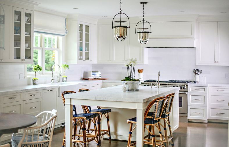 The renewed kitchen was taken from Tuscany to Nantucket with the use of white cabinets, counters and backsplash. The change answered the homeowners’ desires for a light-filled space that honored the home’s exterior architecture. To marry old and new and here and there, the designers used statuary marble on counters, Parisian stools are placed around the island and blue-and-white striped cushions are on the bamboo-like antique chairs by Baker.
