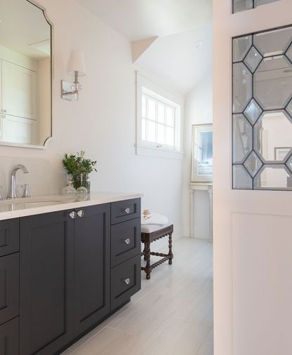 The goal of the downstairs bathroom’s remodel was to up the specialness factor to make it more guest friendly and powder room-like while having the design complement the rest of the home. To attain that objective, the same cabinetry was used as elsewhere but in a dramatic gray, crystal was introduced via the vanity knobs and lighting and plumbing fixtures, and a leaded glass privacy screen was custom made to provide privacy for the toilet.