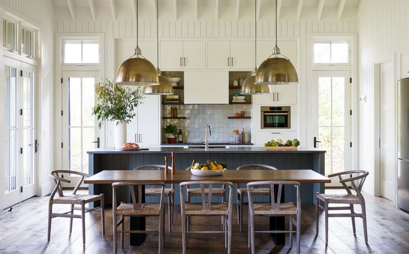 Vaulted ceilings add a sense of spaciousness to the kitchen/dining room when families gather for meals. Wire brushed wide plank oak flooring with custom finish by Natural Creations runs throughout home. Painted Benjamin Moore Gray Garden kitchen island by Traditional Woodcraft. Bosch electric cooktop with Clé Zellige backsplash tiles; freestanding custom stained oak shelving. Shiplap 1x6 walls by Windsor One. Custom hood and cabinetry: Traditional Woodcraft. Oversized Circa Lighting pendants. Custom dining table by Meyer Wells; Four Hands dining chairs.