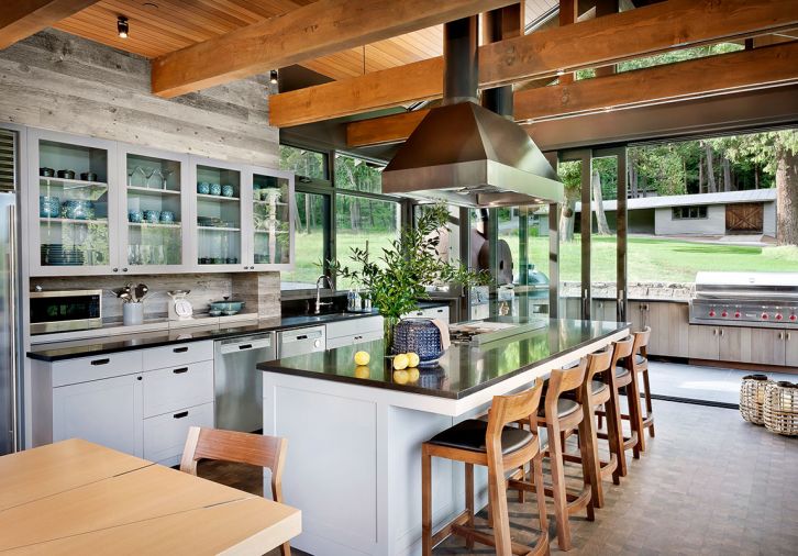 Architects designed the kitchen to flow outdoors through lift and slide Quantum doors to BBQ. Fung specified finishes/detailing. Pioneer Millworks snow fencing trims easy-to-clean painted cabinets – Briarwood (Benjamin Moore). Appliances move forward on custom rolling trays. Absolute Black granite island countertop. Sturdy end grain hemlock flooring from Oregon Lumber & Co. provides texture. Cabinetry and custom dining table with inlaid metal by William Walter Woodworking points to homeowners’ favorite places. Pizza oven doors crafted from marine buoy.