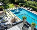 Seen from the second-floor deck seating area with individually precast concrete pavers and custom steel railings, the newly resurfaced pool features new, gradual stepped down entrance alongside spa that gleams in the sunlight. RH Marbella and Aegean outdoor furnishings. Privacy hedge runs length of pool backed by higher shrubs. Retractable canvas roof to capture pine needles protects cabana. Artificial turf looks anything but, with variegated colored blades of “grass.” Turf surrounding individual pavers creates a Greek Key motif.