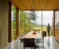 The house is wrapped in a custom window wall system by Quantum Windows and Doors, with deep oak jambs on the interior and metal cladding on the exterior. The David Groppi MISS LED metal pendant lamp by Omar Carraglia is suspended over a table designed by MW Works, paired with chairs purchased by the owner. The teak and blackened steel cabinetry in the kitchen is a handsome divider between the two rooms.