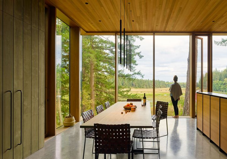 The house is wrapped in a custom window wall system by Quantum Windows and Doors, with deep oak jambs on the interior and metal cladding on the exterior. The David Groppi MISS LED metal pendant lamp by Omar Carraglia is suspended over a table designed by MW Works, paired with chairs purchased by the owner. The teak and blackened steel cabinetry in the kitchen is a handsome divider between the two rooms.