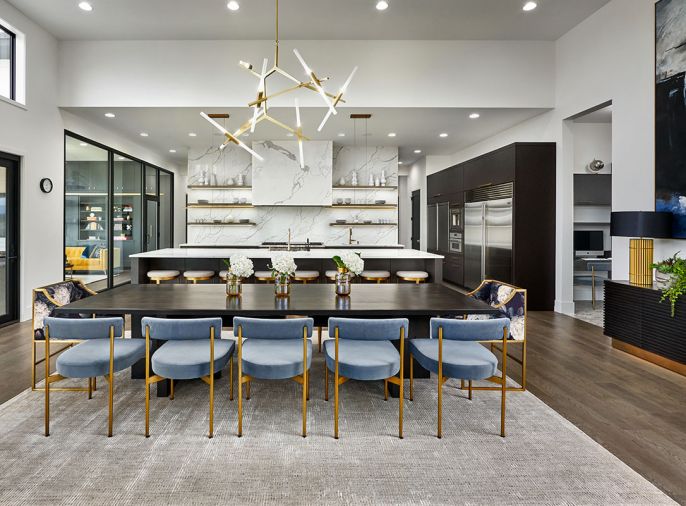 Kitchen island cabinetry by Hayes Cabinets pairs off against custom dining table seating 12. Blue Sunpan dining chairs trimmed in brass. Avenue Lighting chandelier draws eye to Elaborated Stone Design’s custom-crafted Calacatta ESD quartz countertops, range hood and wall. Cleaf laminate finish adds texture to cabinets housing refrigerator and roll-up appliance garage. Built-in office desk and cabinets off right. Marvin windows open view from kitchen into adjacent library/music room and out to river beyond.