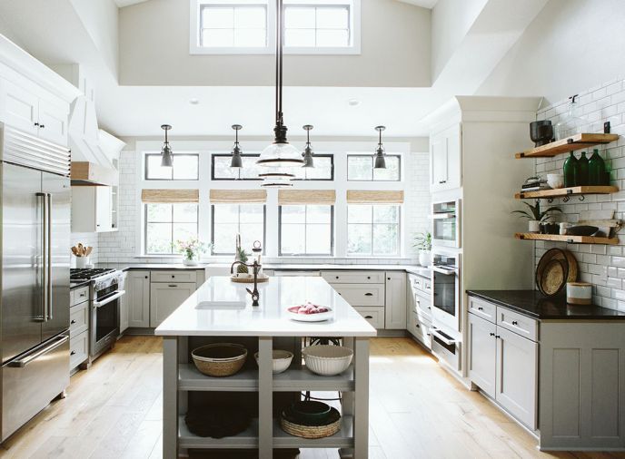 Milgard Black Bean fiberglass windows add both contrast to neutral kitchen and curb appeal to exterior. Globe Lighting furnished Chadwick oil rubbed bronze pendants over sink and clear domed style over island. Awning clerestory windows bring additional light. Argonne Biscuit French Oak flooring grounds white elements. Metal bracketed open wood shelves add farmhouse touch. Custom hood built by Fuller Woodcraft trimmed in French Oak to play off additional wood accents found in bamboo shades. Sit-down island features a Fisher & Paykel dishwasher.