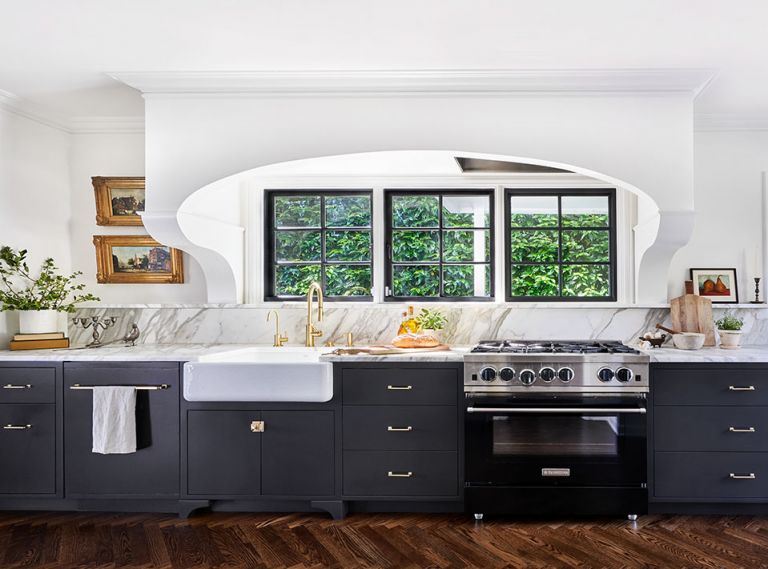 A bank of black cabinets by L&Z Specialties match custom-colored Blue Star range for modern flare. Stunning extra honed Calacatta Daltile installed by Alpha StoneWorks features beautifully crafted 4  linear ledge that draws eye to custom corbels supporting classic arch housing Vent-A-Hood. Rejuvenation Mission unlaquered brass cabinetry hardware is repeated on the more traditional (unseen) white cabinets opposite. Kohler Whitehaven sink from Ferguson. Arlene ditched low-quality cabinets and replaced flipper’s “hideous” wood floors with herringbone oak floors by Champion Hardwoods to better match original floors in adjacent dining room now made more accessible and alluring through a 5  wide opening.