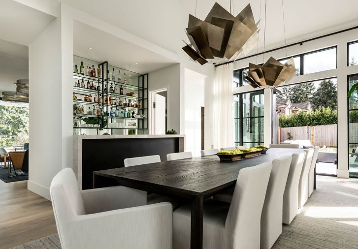 In the dining room, sculptural light fixtures from Curry & Co. are suspended over a table, chairs, and rug from Restoration Hardware. The adjacent bartop is wrapped with a porcelain slab serving counter from CR Floors. The exterior glass double doors open to a sun patio at the front of the home.