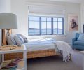 Black-framed Sierra Pacific windows (used throughout) capture the water views, and a chair upholstered in blue fabric cozies a corner. The woven bed frame by Serena & Lily adds textural interest.