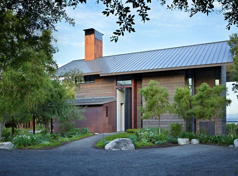 Existing agricultural buildings inspired architect Kirsten Ring Murray to create a steeply pitched metal roof. Horizontal torched spruce siding resembles weathered barnwood. Steel pivot entry door plays off utility room at left, its patina changing with exposure to elements. Island Gardens Company landscaping features a trio of willows creating symmetry with tree at left. Small evergreen Douglas Iris, heather, and creeping thyme are ground cover. Deer fence posts are small steel I-beams.