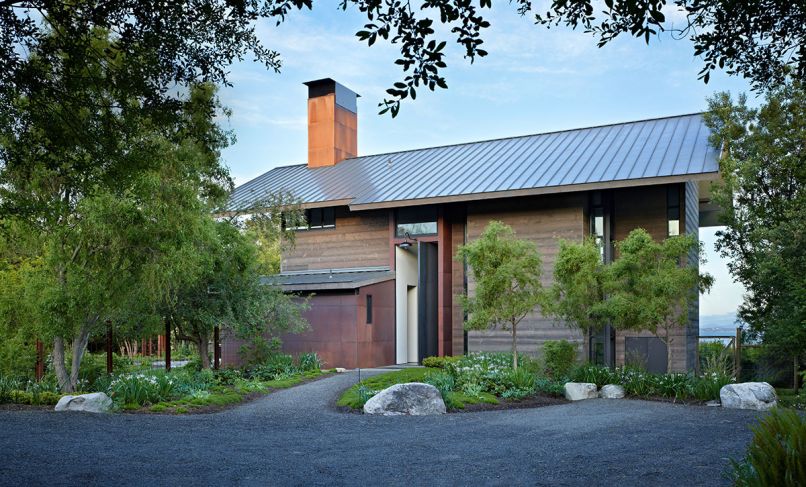 Existing agricultural buildings inspired architect Kirsten Ring Murray to create a steeply pitched metal roof. Horizontal torched spruce siding resembles weathered barnwood. Steel pivot entry door plays off utility room at left, its patina changing with exposure to elements. Island Gardens Company landscaping features a trio of willows creating symmetry with tree at left. Small evergreen Douglas Iris, heather, and creeping thyme are ground cover. Deer fence posts are small steel I-beams.