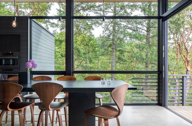 Marvin windows frame views of all aspects of the site, from water to forest. The kitchen island is topped in Absolute Black granite in a silk finish, with counter-height Cherner stools beneath. The pendant lights above are suspended from a track, both by Bruck Lighting.