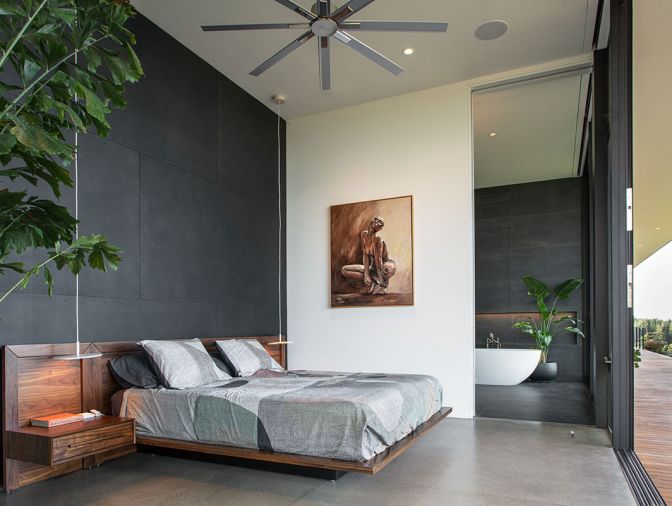 In the primary suite, the blackened steel casework continues behind the bed. Floor to ceiling TruStile pocket door from Portland Millwork is cleverly concealed between bedroom and bath. Large expanses of glass connect with the yard and view, but window coverings by Mari Design ensure privacy and shade when needed. Photography © Peter Eckert