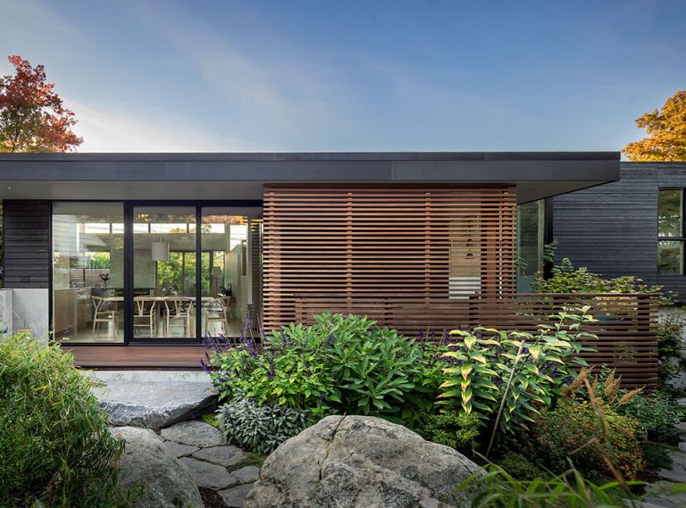 Grounded, organic sensibilities are reflected in the gardens and massing of the house. Natural stones from Marenakos Rock Center crop up against the split-levelled house trimmed in Blackened Cedar Open Rainscreen Siding from LS Cedar. E&H Construction created the clear cedar sliding screen on site. Chin designed the solid maple topped dining table left with kitchen and living room beyond. At right, up a half-story, is a guest bedroom.