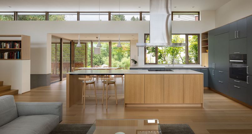 The open plan places kitchen and living areas back-to-back with White Oak flooring that ground everything. Kitchen island features Caesarstone countertop, GE Cooktop, Proline Island Range Hood, and Mattiazzi Radice stools beneath Foscarini Aplomb pendants. Chin’s custom dining table lit by Leucos Celine pendant. Custom cabinetry by Space Theory Henrybuilt Cabinets with Miele oven located in contrasting dark cabinets right. Julien sink with Franke Pescara faucet. At left, custom bookshelves atop staircase mimic the clerestory window design.