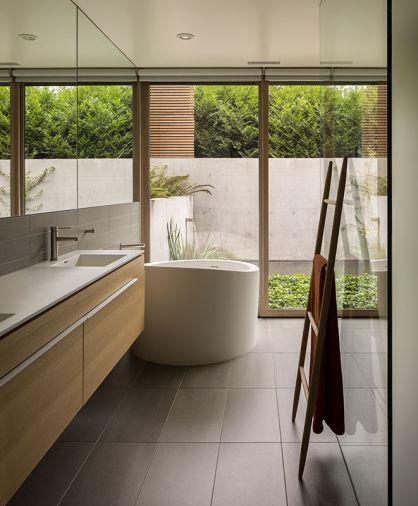 Primary Bath with Aquatica True Ofuro Japanese soaking tub recessed into floor for easier access. Barnes Wood custom vanity cabinetry with MTI sink bowls seamlessly integrated with countertop. Graff faucet. MOSA Core Collection Solids floor and wall tile. Water feature beyond drowns traffic noise.