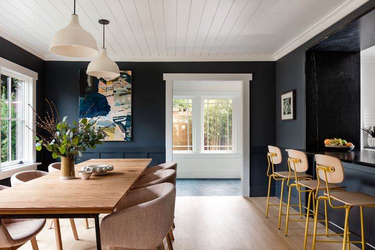 Dark walls in the dining area feature Benjamin Moore paint in Graphite, a blue that shifts from a deep marine hue to black as light moves through the space. A new doorway offers access to a sunporch the homeowners use as a drop zone and a new cocktail prep and bar storage area. The play of blacks and whites throughout acts as a neutral backdrop that’s anything but bland. Pops of subtle color find their way into accents and fixtures, including gold barstools from Grand Rapids Chair and pink upholstered dining chairs by BluDot. The enlarged window allows ample natural light to wash through to the kitchen area and garden views.